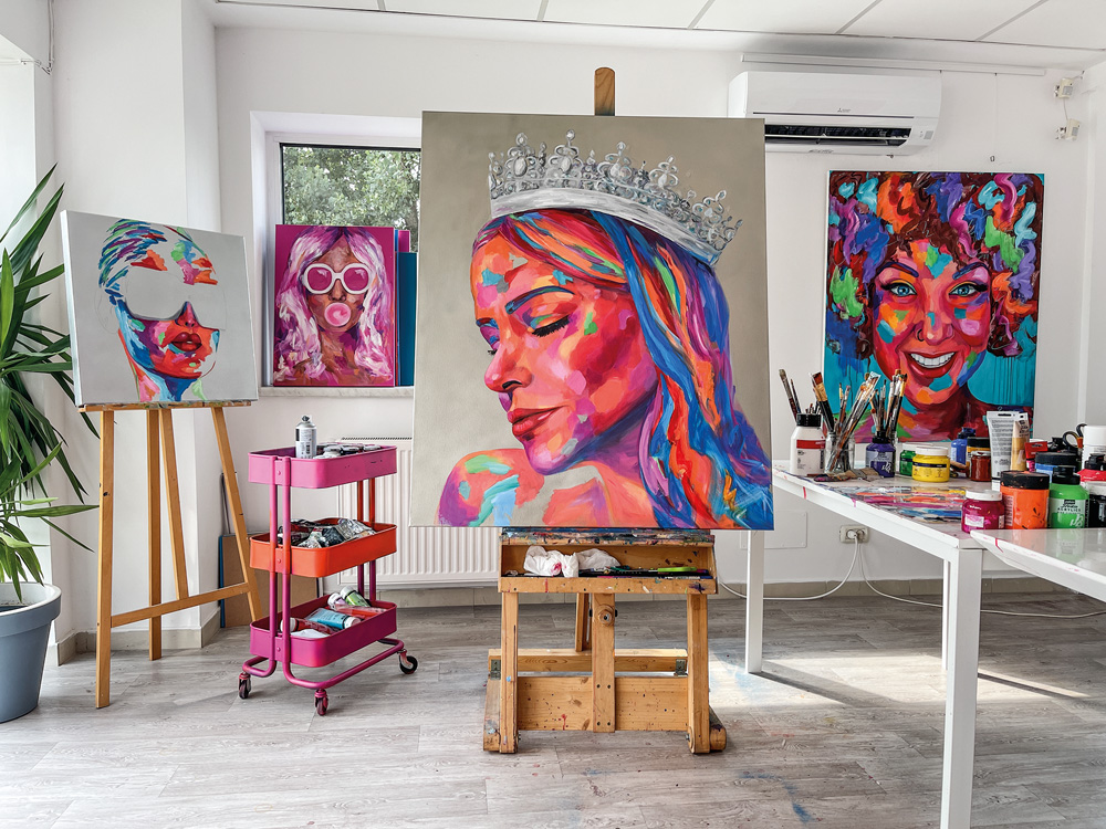 Obra de Ramona Pintea aparece erguida em um ateliê. O quadro exibe o rosto de Natalia Martins em versão colorida (rosa, azul e roxo) com uma coroa na cabeça).