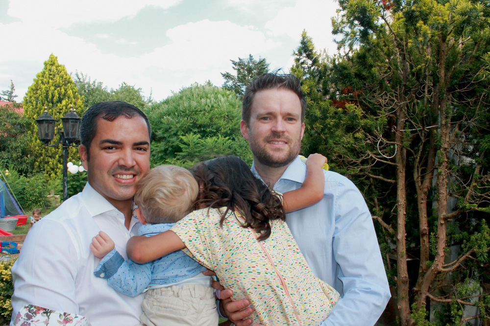 Rodrigo Santana e Patrick Hohmann (ARQUIVO PESSOAL).JPG A imagem mostra Rodrigo e Hohmann segurando seus dois filhos, que estão de costas para a câmera. O casal de pais está sorrindo para foto enquanto seus filhos os abraçam na altura dos ombros.