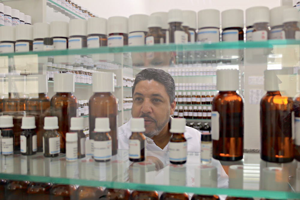 Samuel Moraes fotografado por detrás de prateleira translúcida de frascos de fragrâncias analisando-os
