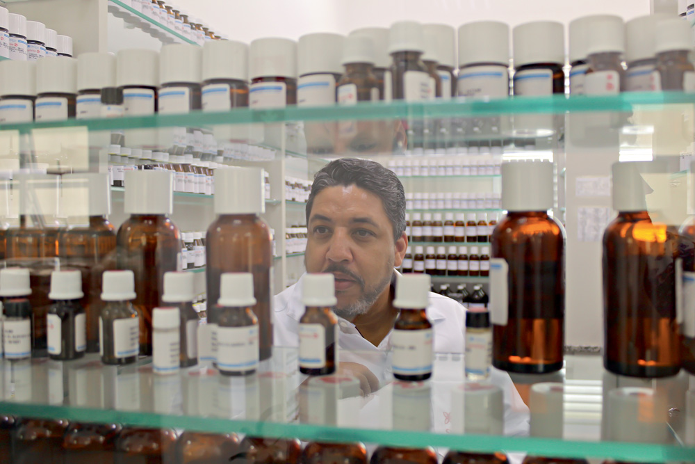 Samuel-Moraes-perfumistas-rotina-fragrâncias-perfumes Samuel Moraes fotografado por detrás de prateleira translúcida de frascos de fragrâncias analisando-os