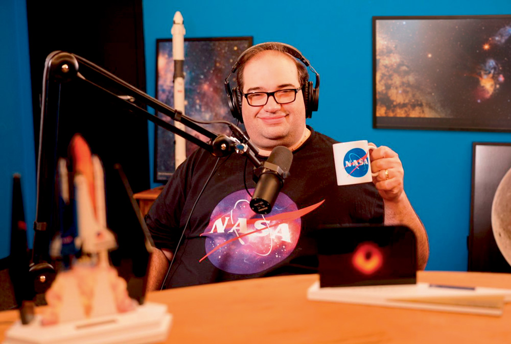 Sérgio Sacani aparece erguendo caneca com logotipo da NASA em frente a microfone de estúdio e fones de ouvido. Veste camiseta preta também com logo da NASA.