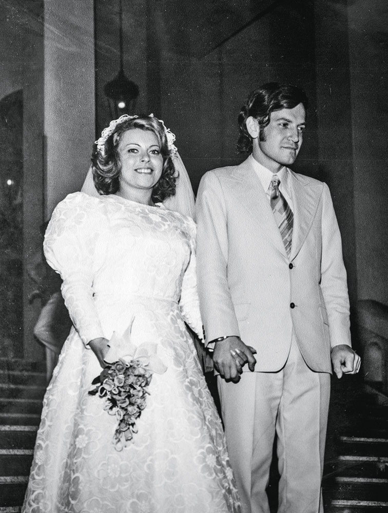 foto em preto e branco de vera e jeri na cerimônia de casamento. eles estão de mãos dadas e vera está com seu vestido de noiva e com um buquê de flores e jeri está com seu terno