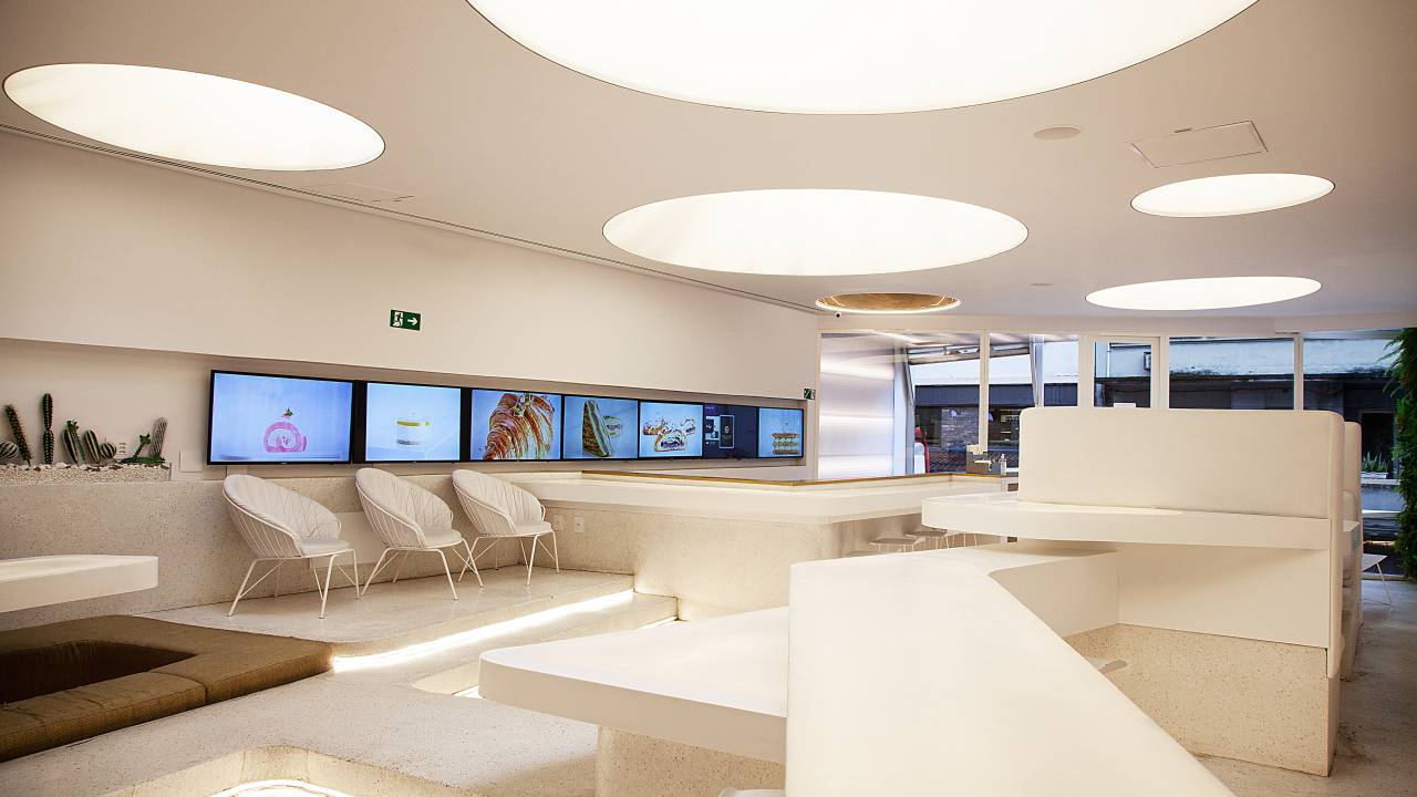 Salão da We Coffee, com paredes, teto e móveis em branco e TVs exibindo salgados à esquerda.