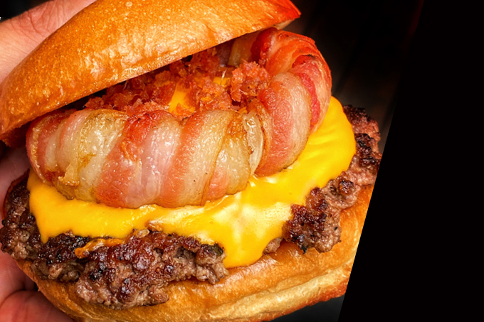 No canto esquerdo hambúrguer com zoom montado com pão, carne, queijo e anel de bacon.
