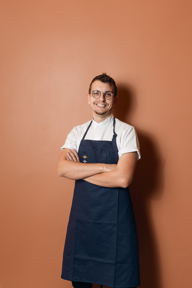 Chef Bruno Hoffman de avental azul e camiseta branca em frente à parede salmão.