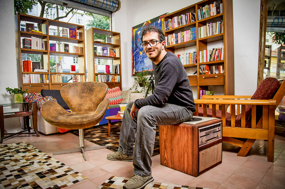 bruno eliezer sentado em banqueta fofa estilizada em sua livraria, ponta de lança. ele está com um braço apoiado em uma das pernas e com o tronco levemente arqueado