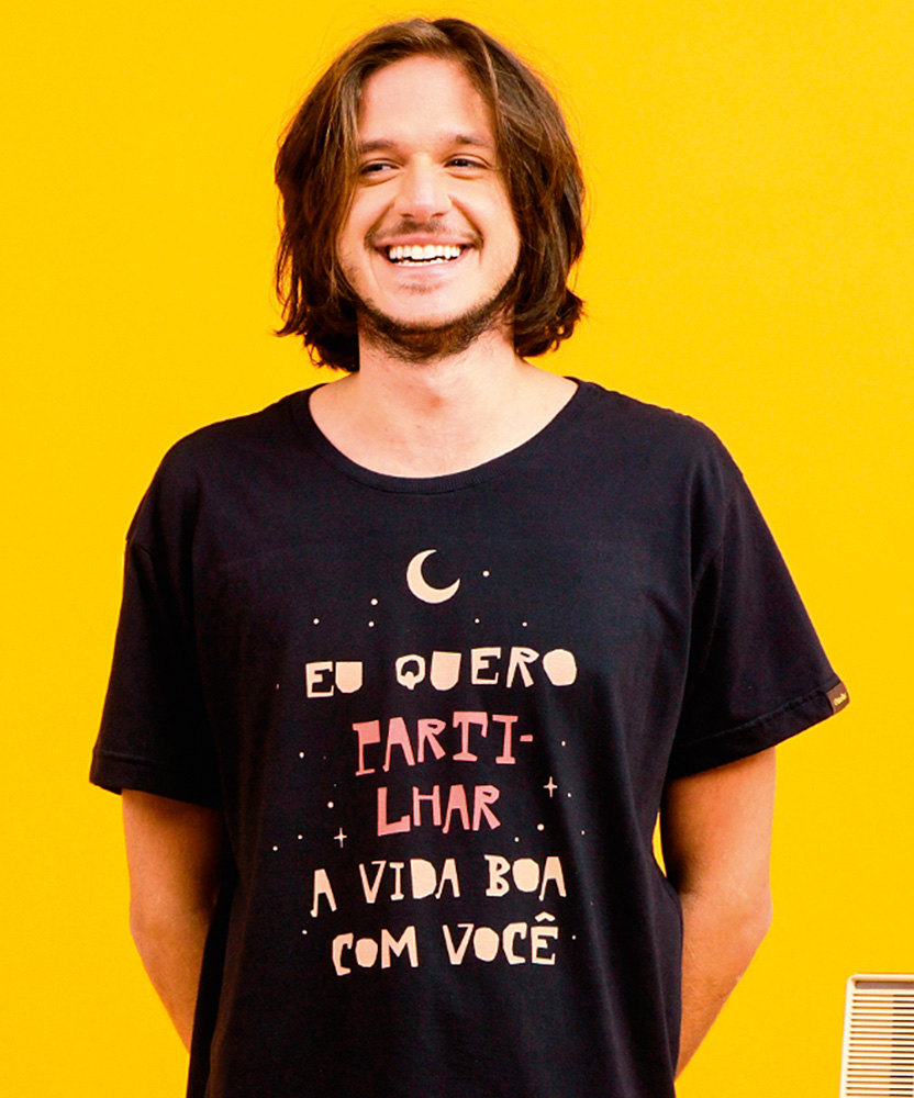 Um homem sorri em um fundo amarelo vestindo uma camiseta preta com a frase 