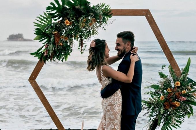 tati e gabriel abraçados com roupa de casamento sorrindo um para o outro. eles estão em um pedaço de grama a frente do mar, com um altar com plantas