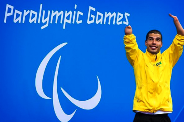 Daniel aparece em frente de parede onde pode-se ler: Paralympic Games. Ele está com os braços erguidos, sorri e usa jaqueta do Brasil