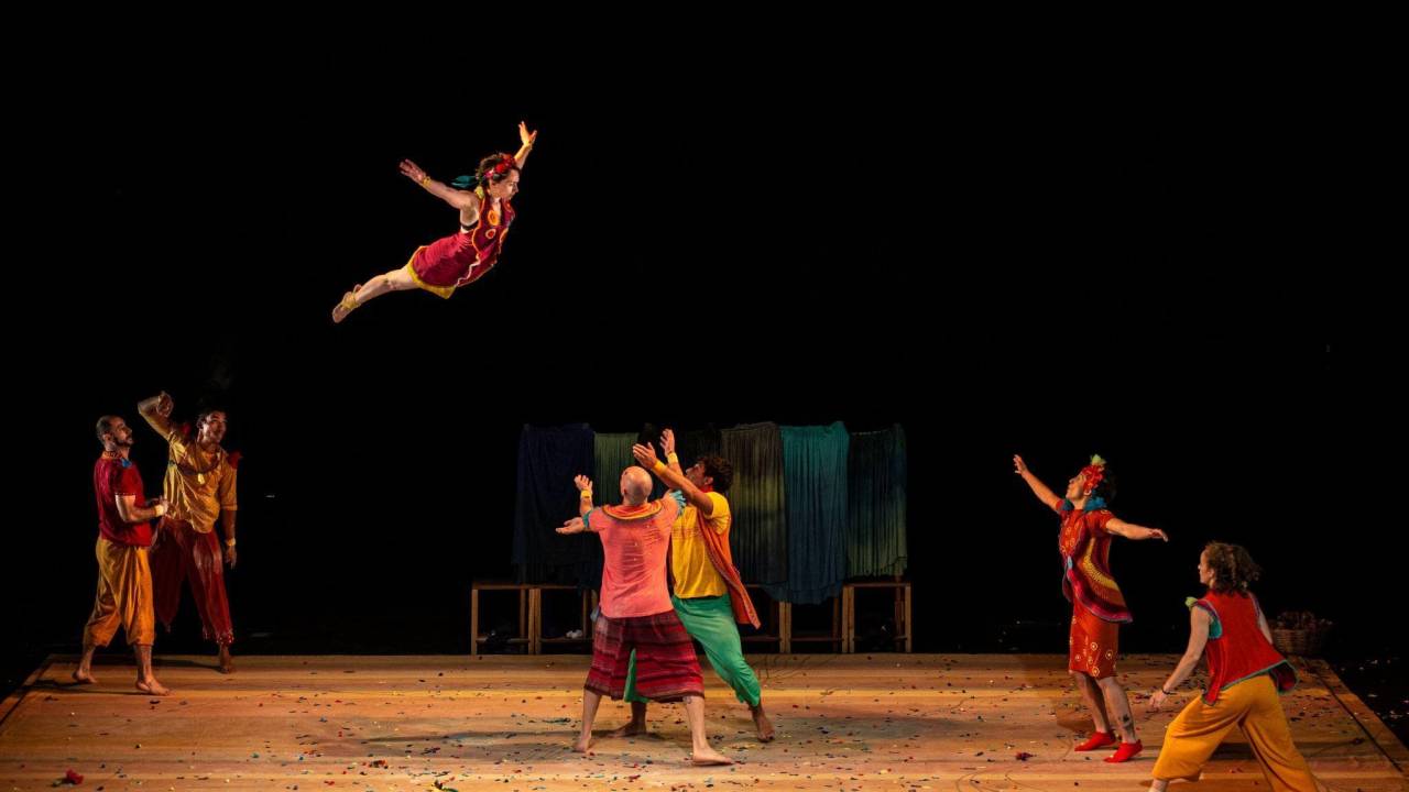 Em um palco de teatro, uma mulher está saltando bem alto enquanto atores estão em posição de segurá-la. Eles estão com roupas de circo