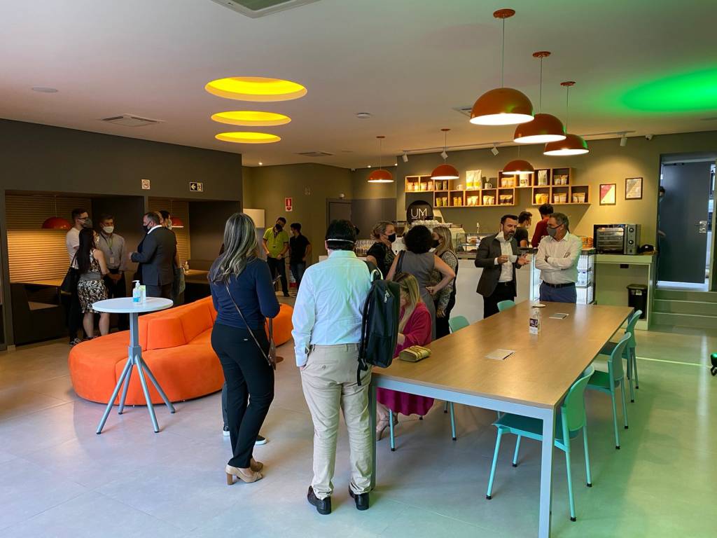 Salão da unidade da Faria Lima do Um Coffee Co. com pessoas em pé, cabines de mesas à esquerda e uma mesa retangular comprida à direita.