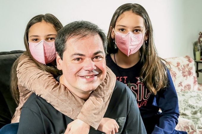 Luis Eduardo e as filhas Marina e Rachel sorrindo para a foto. As meninas estão de máscara e luis está sem. uma das meninas abraça luis por trás, cruzando os braços