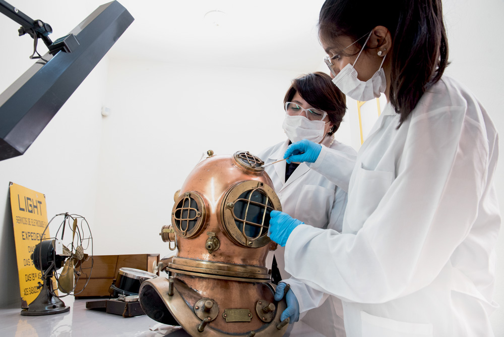 duas funcionárias do museu da energia limpando um escafandro com grandes cotonetes e vestidas adequadamente, com avental, luvas e máscara de proteção contra a Covid-19