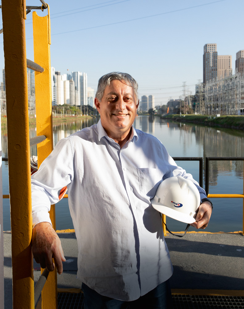 itamar sorrindo para a foto em plataforma sobre o rio pinheiros, apoiado em uma escada amarela e segurando o capacete branco no outro braço. ao fundo, o rio e prédios da cidade