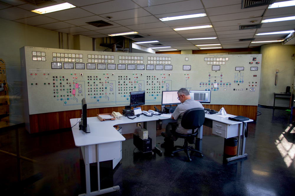 sala de controle da usina de traição, com um painel suspenso com muitos botões e telas. à frente do painel, tem um técnico em sua mesa