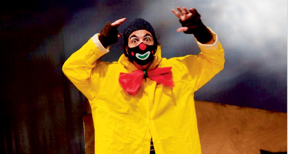 Um palhaço está com uma capa de chuva amarela e levanta os braços, espantado, num palco de teatro