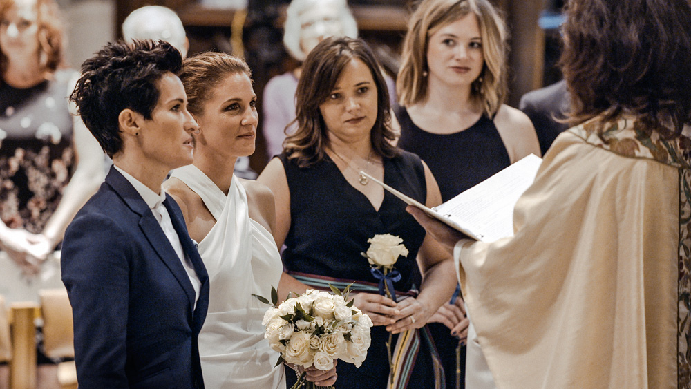 A imagem mostra duas mulheres em um altar, se casando. Uma está de vestido, segurando um buquê, outra está de terno. À frente delas, uma outra mulher conduzindo a cerimônia.