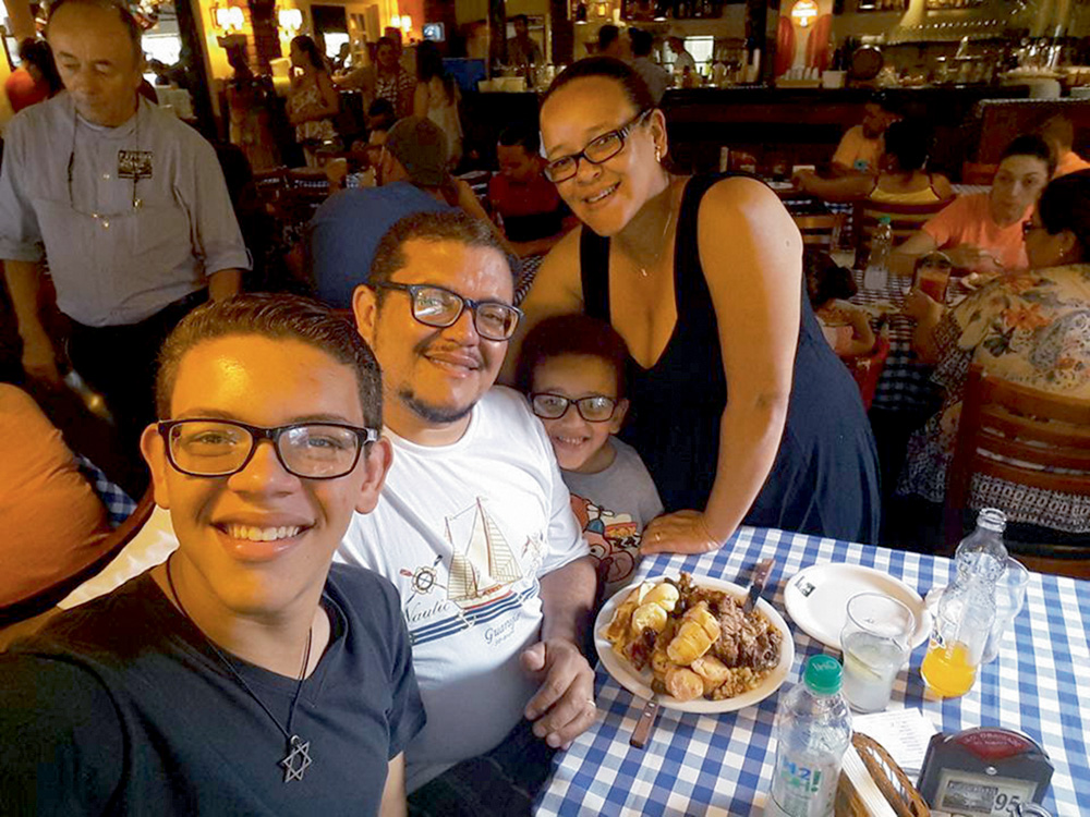 Ryan Henrique e Ryan Victor com os pais, Ecio e Fernanda, sorrindo para selfie em restaurante. ryan henrique, que estão tirando a foto, está de pé assim como fernanda e ryan victor. ecio está sentado, com um prato cheio de comida à sua frente
