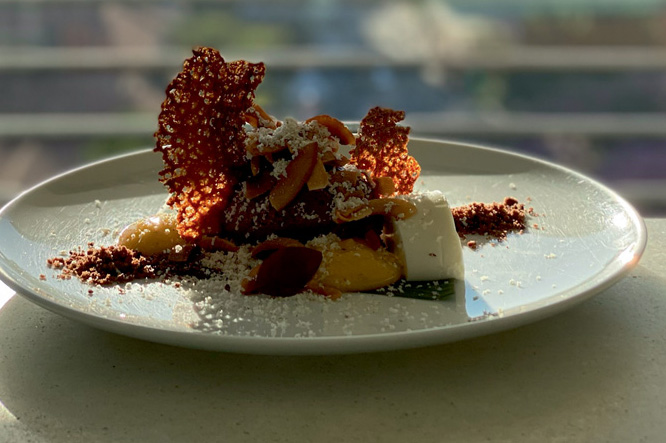 Cheesecake coberta por calda amarelada, tela caramelo e castanhas posicionada sobre uma mesa de frente para a parede de vidro do restaurante Esther Rooftop, com vista para a Praça da República.