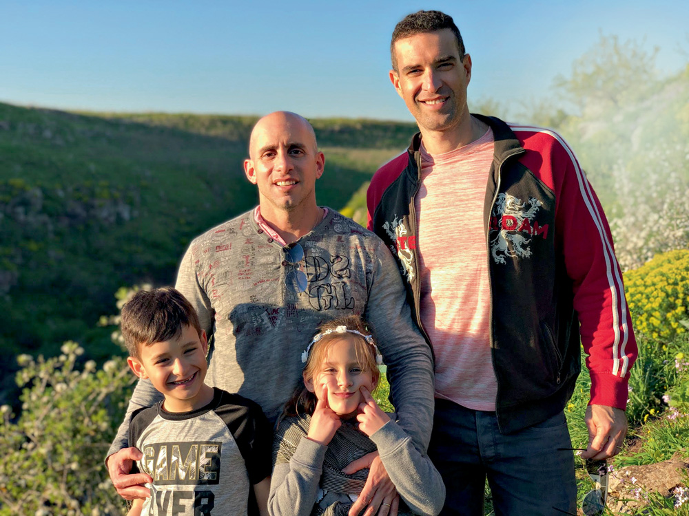 Roy Rosenblatt-Nir Ronen A imagem mostra Roy e Ronen, os dois de pé em um campo aberto e verde. Na frente deles, sendo abraçados por Roy, estão duas crianças da altura da barriga de Roy, sorrindo para a câmera.