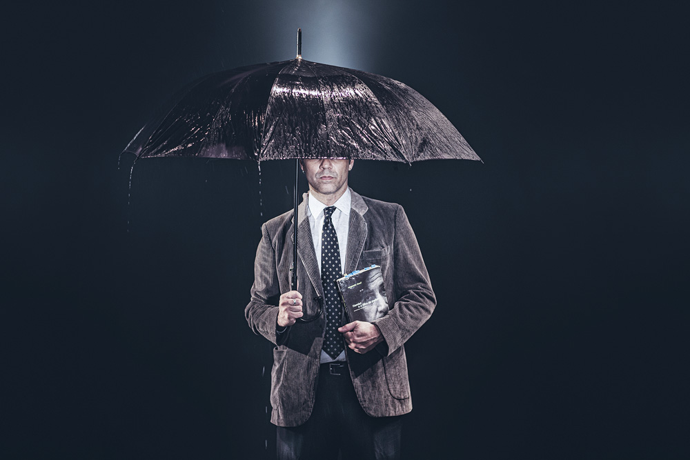 A imagem mostra um homem de roupa social, terno e gravata, segurando um livro e um guarda-chuva molhado. Ele está em um fundo preto e não é possível ver seu olhos.