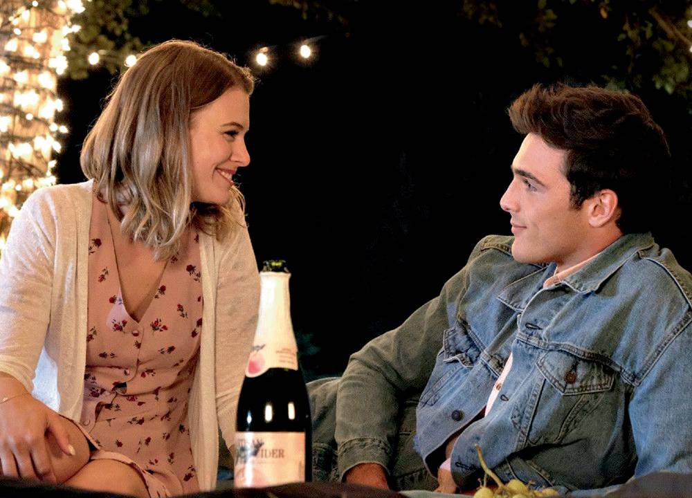 A imagem mostra um casal de um homem e uma mulher. Eles estão sentado em uma mesa, a noite, olhando um para o olho do outro, sorrindo.
