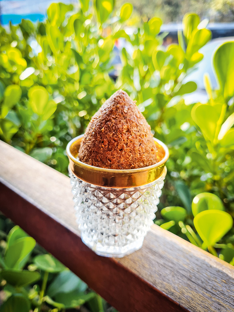 Copinho de cristal com borda dourada com quibe frito dentro apoiado em corrimão de madeira com plantas atrás.