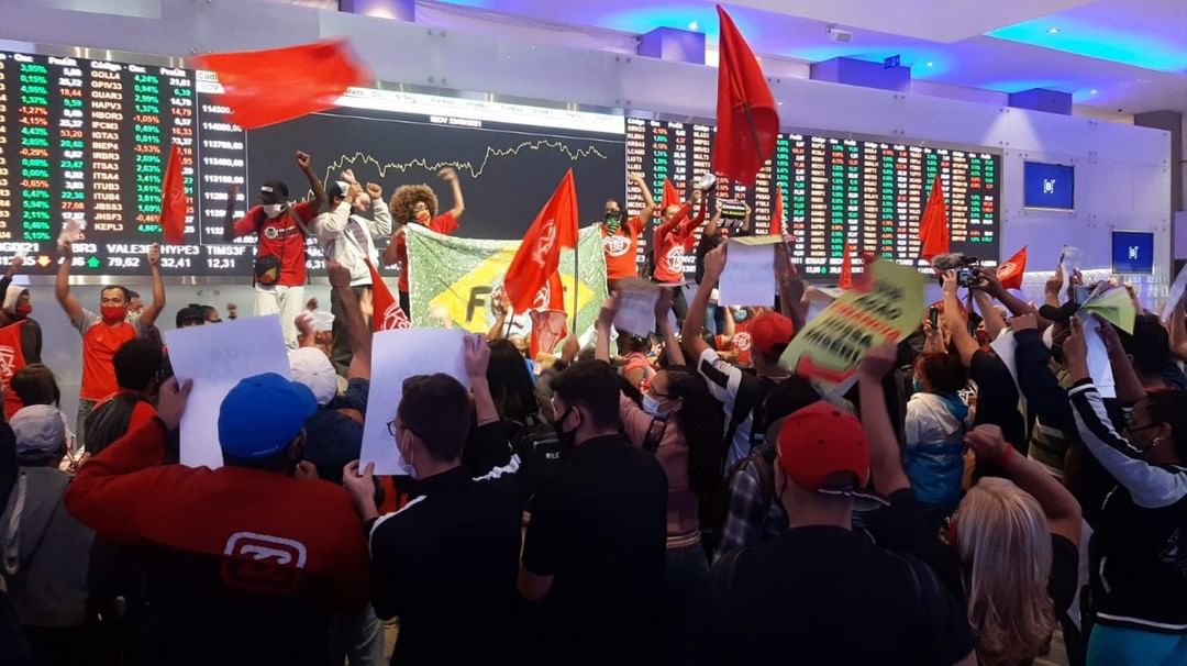 Imagem mostra dezenas de pessoas carregando bandeiras do MTST, com bandeira do Brasil ao fundo, na frente de telão de cotações da Bolsa de Valores, a B3