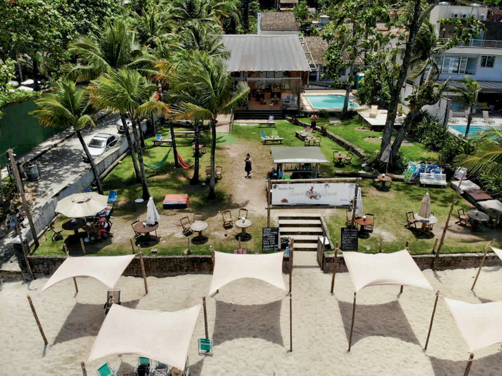 Foto aérea mostra uma área de um bar restaurante à beira-mar. Tem quiosques, piscina e um grande gramado