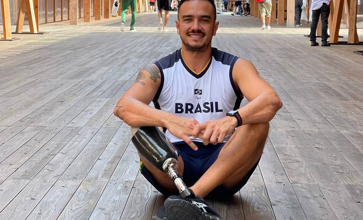 Um homem com blusa do Brasil está sentado no chão de uma passagem pública. Ele está sentado com as pernas cruzadas, uma das pernas é amputada e mecânica