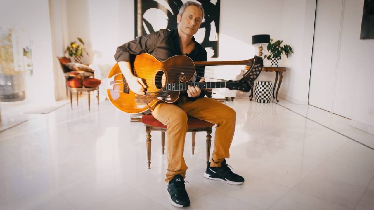 O produtor musical Apollo Nove posa sentado em sala ampla segurando guitarra. Veste calça bege e encara a câmera.