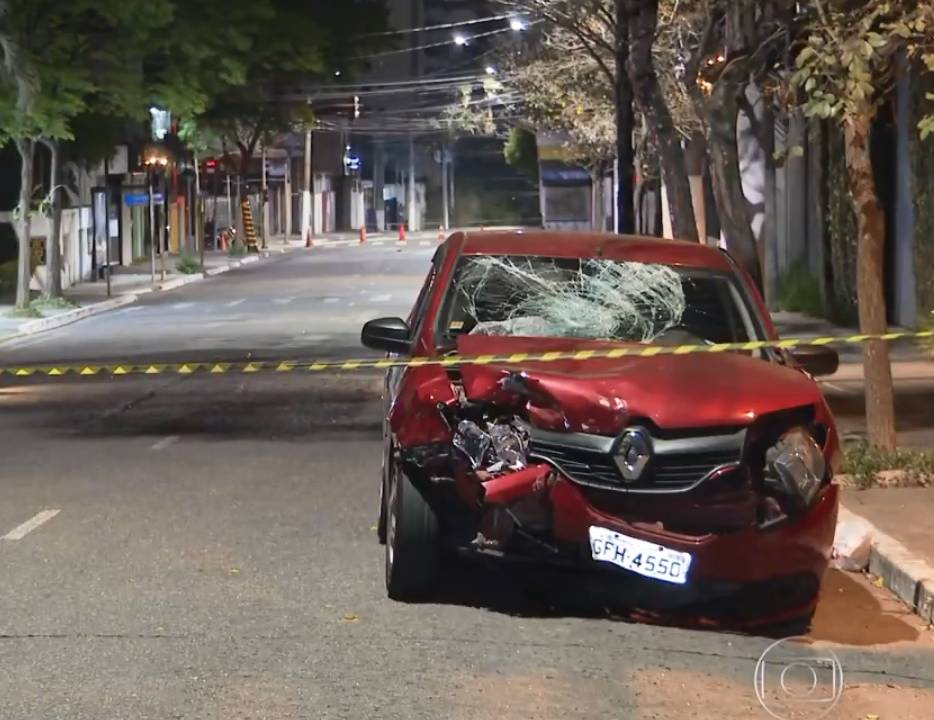 A imagem mostra um carro vermelho, com o vidro do para-brisa amassado e estilhaçado. A parte de frente do carro, faróis, estão desconfigurados.