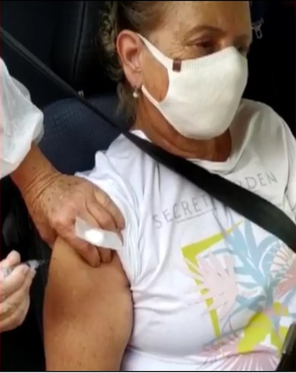 Idosa com máscara está no carro com cinto de segurança enquanto uma mão segura a seringa de vacinação.