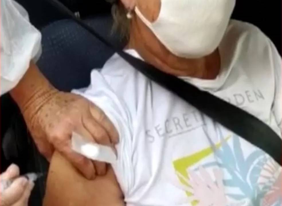 Idosa com máscara está no carro com cinto de segurança enquanto uma mão segura a seringa de vacinação.
