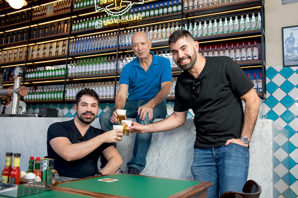 A imagem mostra Joarez, sentado no balcão do bar, e seus dos filhos ao lado, um sentado em uma cadeira e outro de pé, brindando copos cheios de chopp.