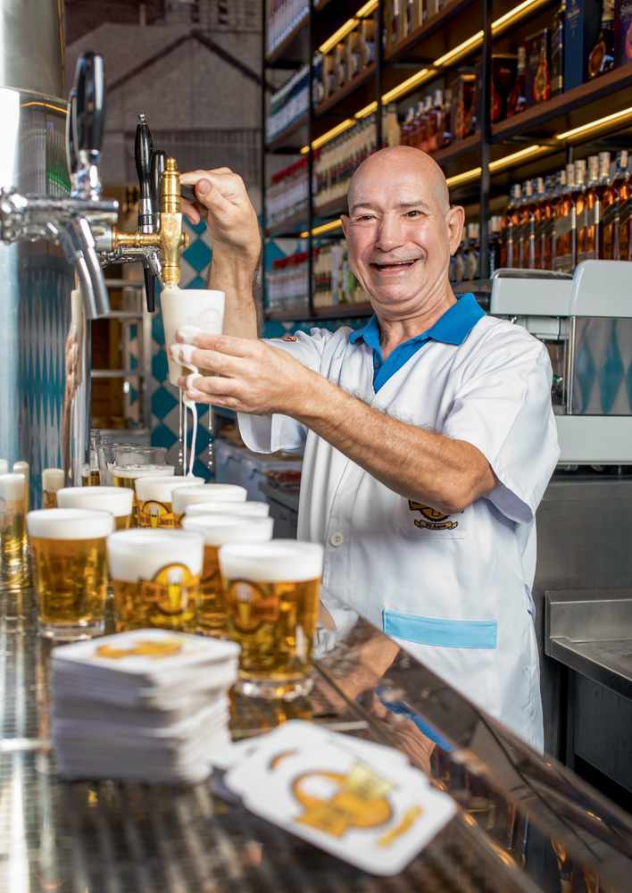 A imagem mostra Joarez enchendo um copo em uma chopeira enquanto o líquido da bebida escorre para fora do objeto. Ele está de avental e sorrindo.