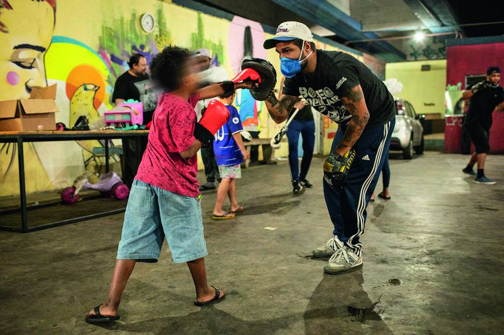 A imagem mostra uma criança, dentro de uma sala, socando um luva de proteção que está na mão de um adulto, que o treina.