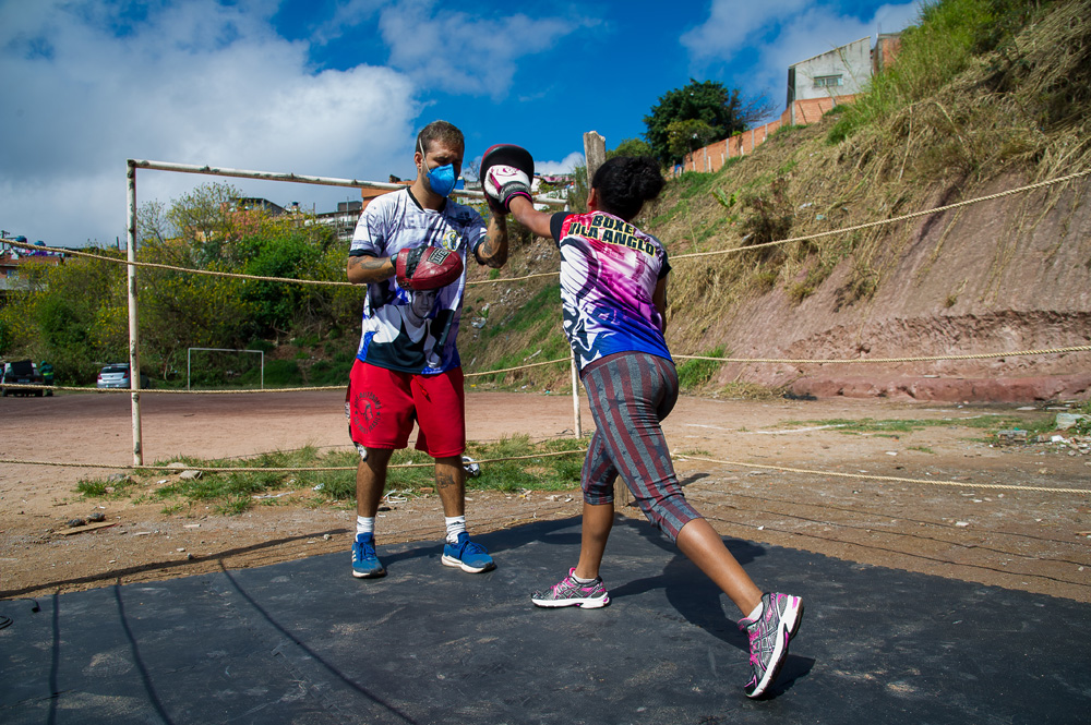 A imagem mostra um homem, de máscara, com uma luva de treino de boxe recebendo um soco justamente dela por uma criança, jovem. Eles estão em um ringue improvisado e ambos de máscara.