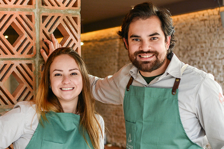 Giovanna Perrone e Rodrigo Aguiar posam lado a lado em frente ao seu novo restaurante, o Casa Rios. Ambos usam avental de cor verde com o nome do restaurante estampado próximo ao peito.