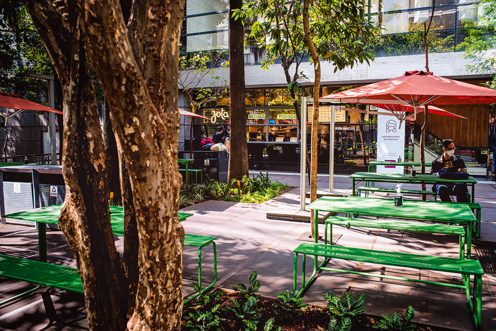 Praça na lateral do Shopping Cidade São Paulo com mesas metálicas de cor verde e guarda-sóis de cor vermelha em meio à pequenas árvores de jabuticaba