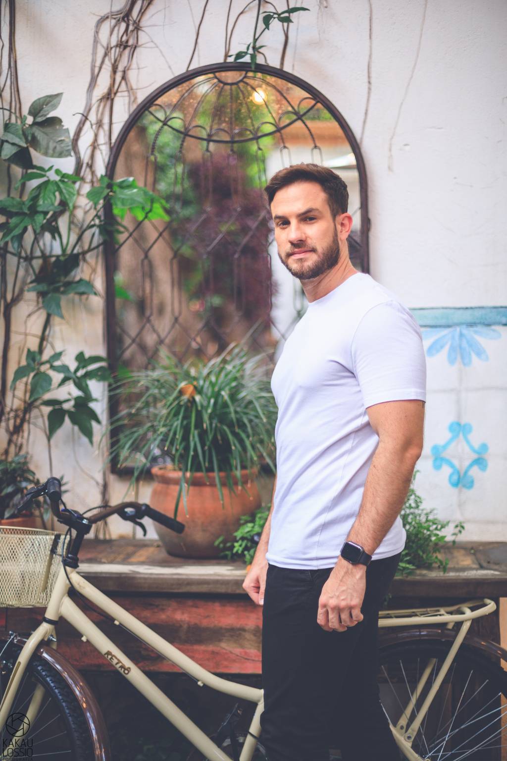Daniel Adjuto (Kakau Lossio) 2 Daniel Adjuto posa de pé, de lado, olhando para a câmera. Está ao lado de plantas e uma bicicleta vintage. Veste calça escura e camiseta branca.