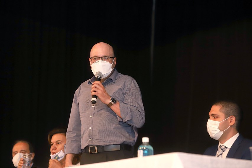 A imagem mostra Dario Saadi, com um microfone, de pé em frente à uma mesa usando uma máscara Pff2