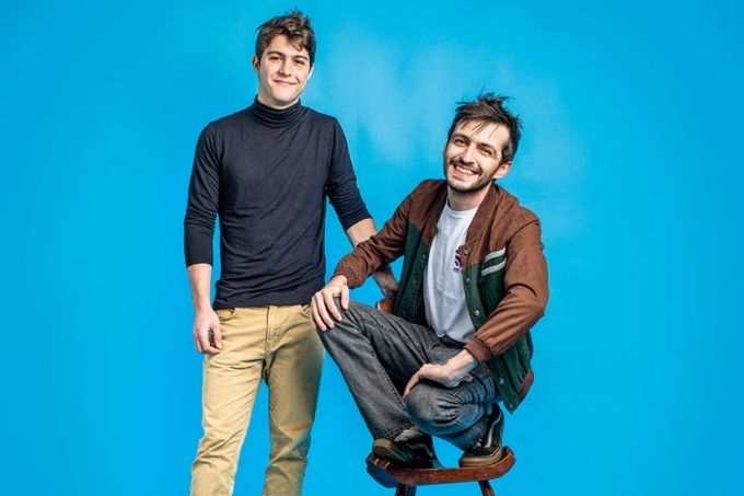 Em um fundo azul, dois homens posam sorrindo para a câmera. Estão em um estúdio de fotos. Um deles está sentado em um banquinho e o outro ao lado dele de pé