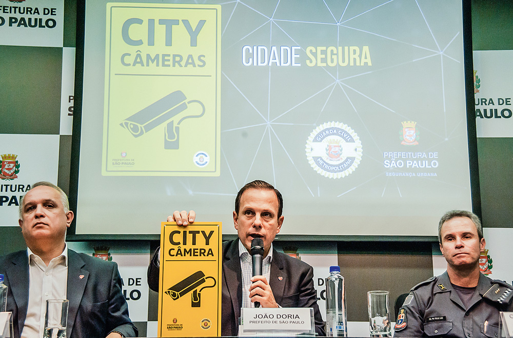 joão doria sentado discursando fazendo anúncio do City câmeras, segurando placa do projeto e com slide projetado atrás dele