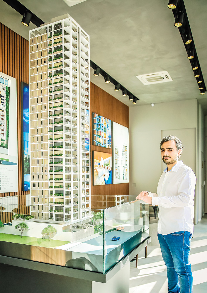 Um homem de camisa social posa ao lado de uma maquete de um prédio de luxo em uma sala comercial