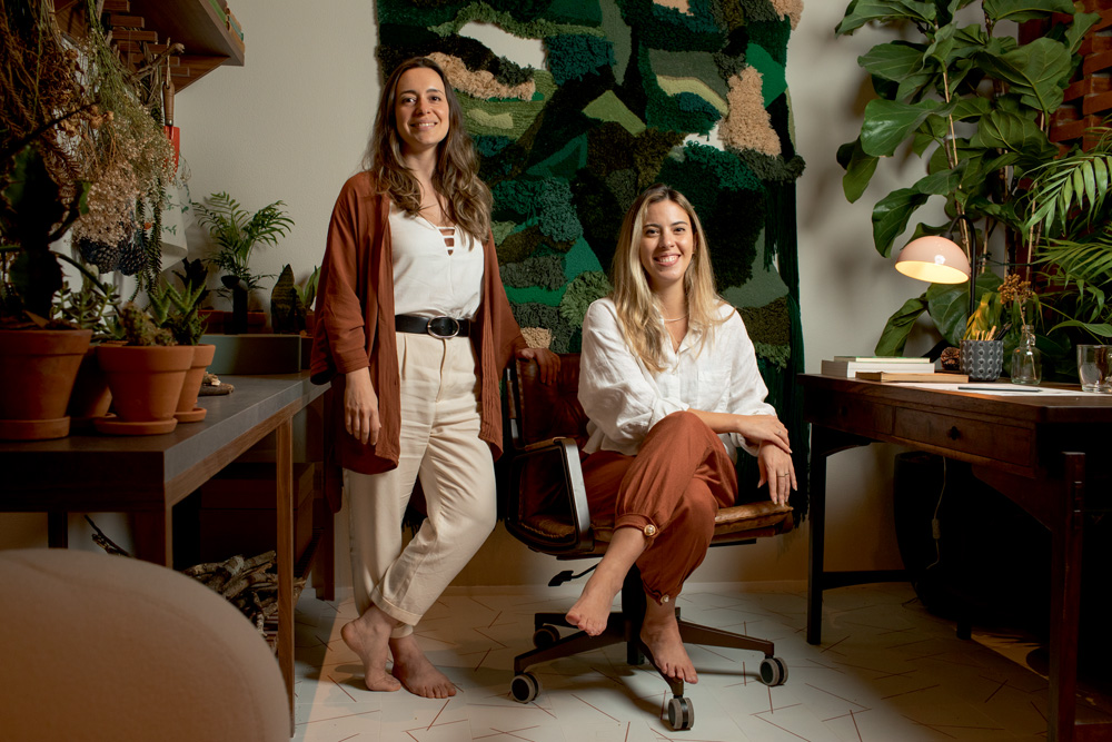 A imagem mostra Fabiana Silveira e Patricia De Palma, a primeira sentada e a segunda em pé, sorrindo para a câmera em seu ambiente.