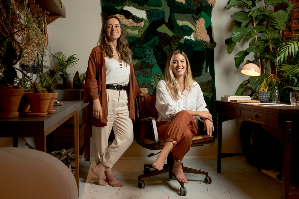 Fabiana Silveira e Patricia De Palma casacor A imagem mostra Fabiana Silveira e Patricia De Palma, a primeira sentada e a segunda em pé, sorrindo para a câmera em seu ambiente.
