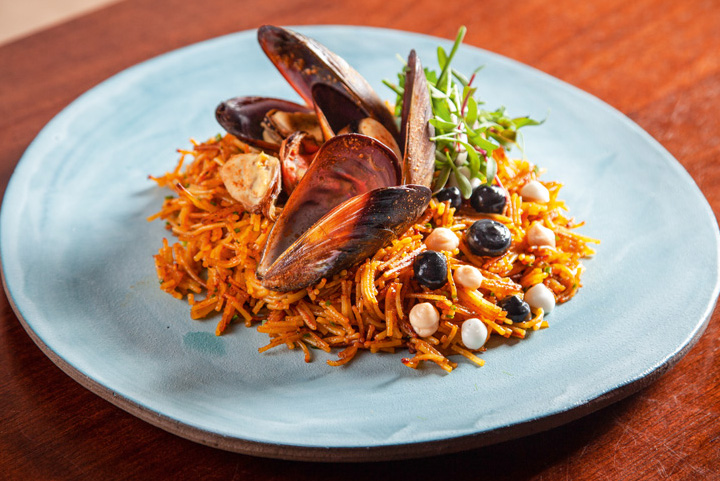 Em um prato raso de cerâmica, é servida a fideuá junto de mexilhões e folhas por cima do macarrão de tipo capellini