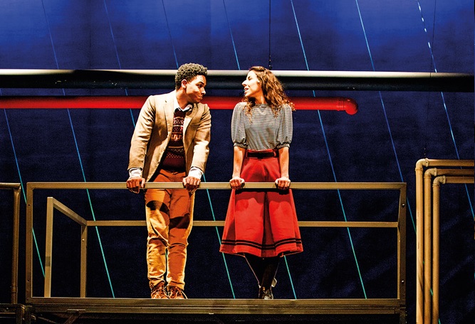 Dois jovens (menina branca e menino negro) estão conversando em uma cena de peça em palco de teatro. Eles estão em uma estrutura como uma escada alta