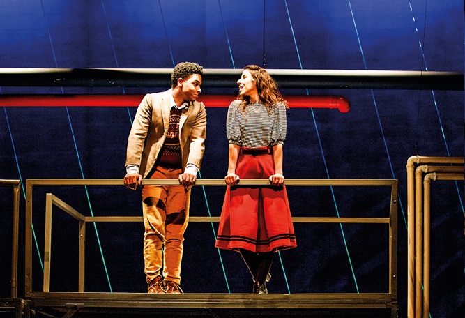 Dois jovens (menina branca e menino negro) estão conversando em uma cena de peça em palco de teatro. Eles estão em uma estrutura como uma escada alta